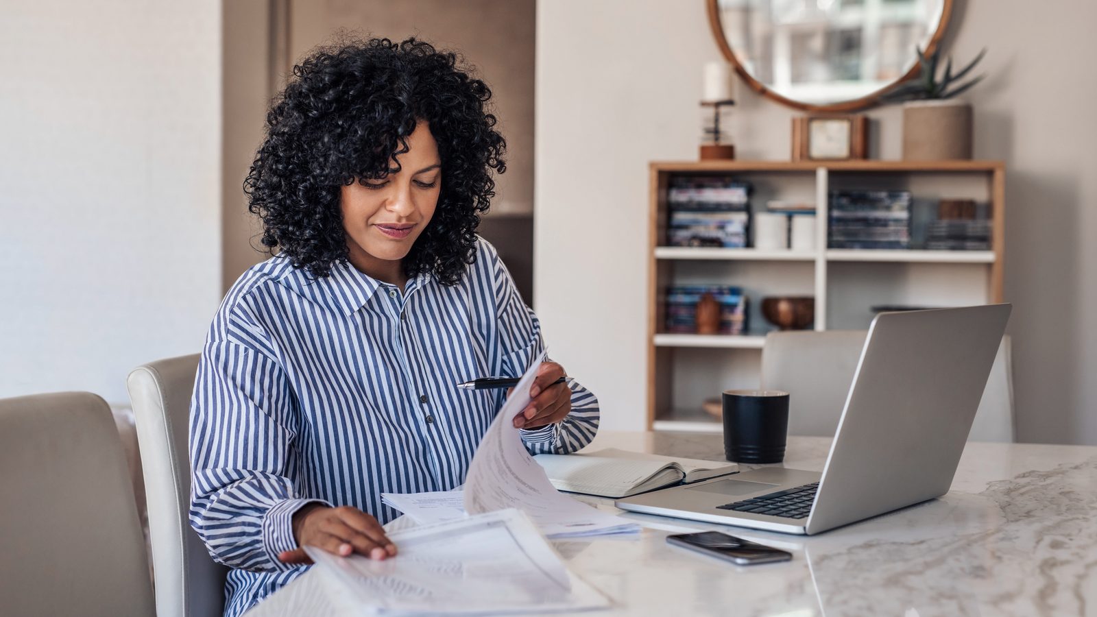 A woman at home reading through paperwork, a laptop open beside her